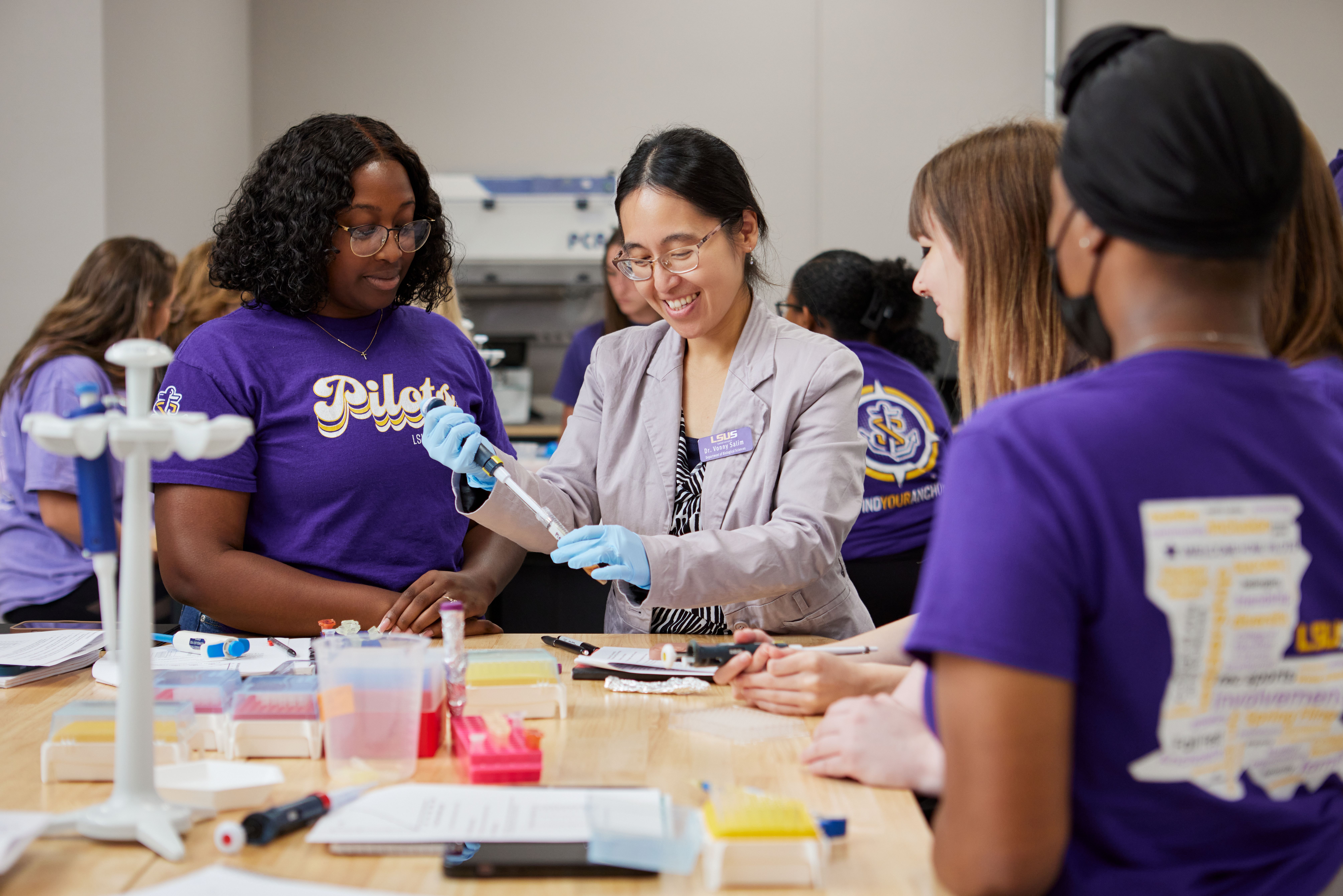 LSUS Professor Vonny Salim, PhD happily demonstrates how to use the modern pipette to fill the syringe with fluid for 4 female students gathered around a workstation in the microbiology lab. The table is strewn with various scientific instruments. The background shows other classmates gathered around other lab workstations. 